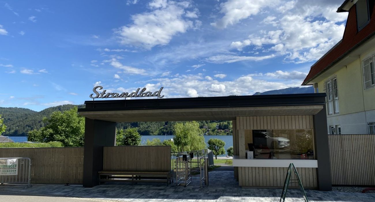 Der neue Eingangsbereich des Strandbad Seeboden. Ein aus Holz gefertigtes kleines Gebäude mit der Aufschrift Strandbad. Zuätzlich gibt es ein Drehkreuz für die Kontrolle, nebenan ist der Kassenbereich hinter einen großen Glasscheibe. 