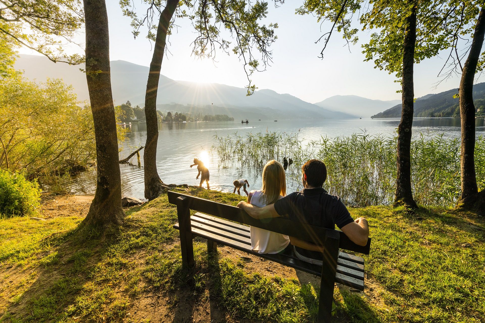 Zwei Personen auf einer Bank im Klingerparkt, die am Ufer des Millstätter See sitzen und ihren Kindern beim Spielen zusehen. Fotorechte-/Archiv Millstatter See Tourismus GmbH Fotograf Gert Perauer.jpg