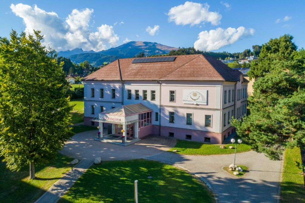 Das Gebäude der VS Seeboden ist in Rosatönen gehalten, mit einem verglasten Eingang an der Front ist eine große Sonnenuhr angebracht.