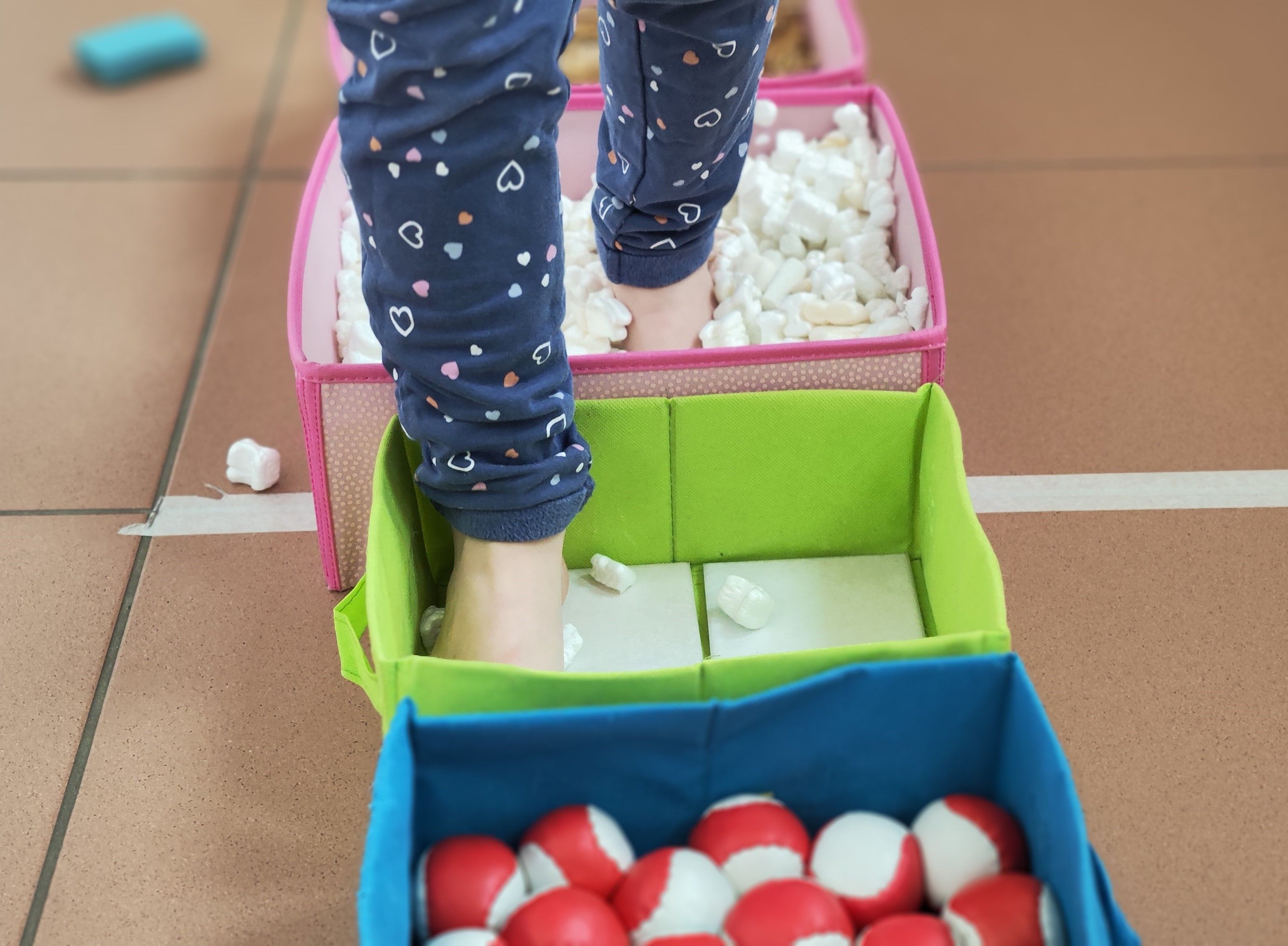 Kinderfüße die durch einen Barfußweg stapfen und unterschiedliche Texturen von Bällen, Stoff oder Verpackungsmaterial erfühlen.