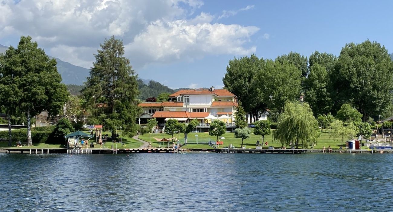 Der Blick auf das Strandbad vom See aus. Eine schöne Liegewiese mit zwei Stegen im Wasser. Im hinteren Bereich ein gelbes Haus in dem sich auch die Gastronomie befindet. 