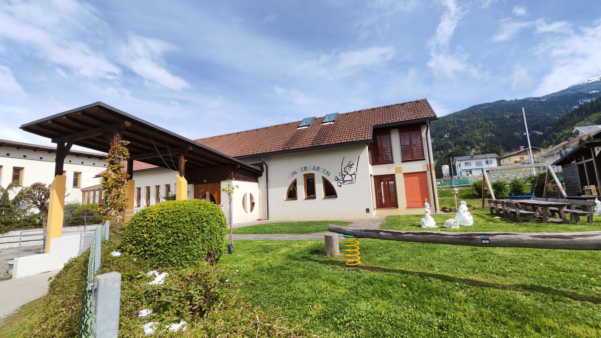 Der Kindergarten Treffling besteht aus einem Gebäude, mit weißer Fassade – die Fenster sind kreisförmig und das Logo wurde auf die Fassade gemalt.