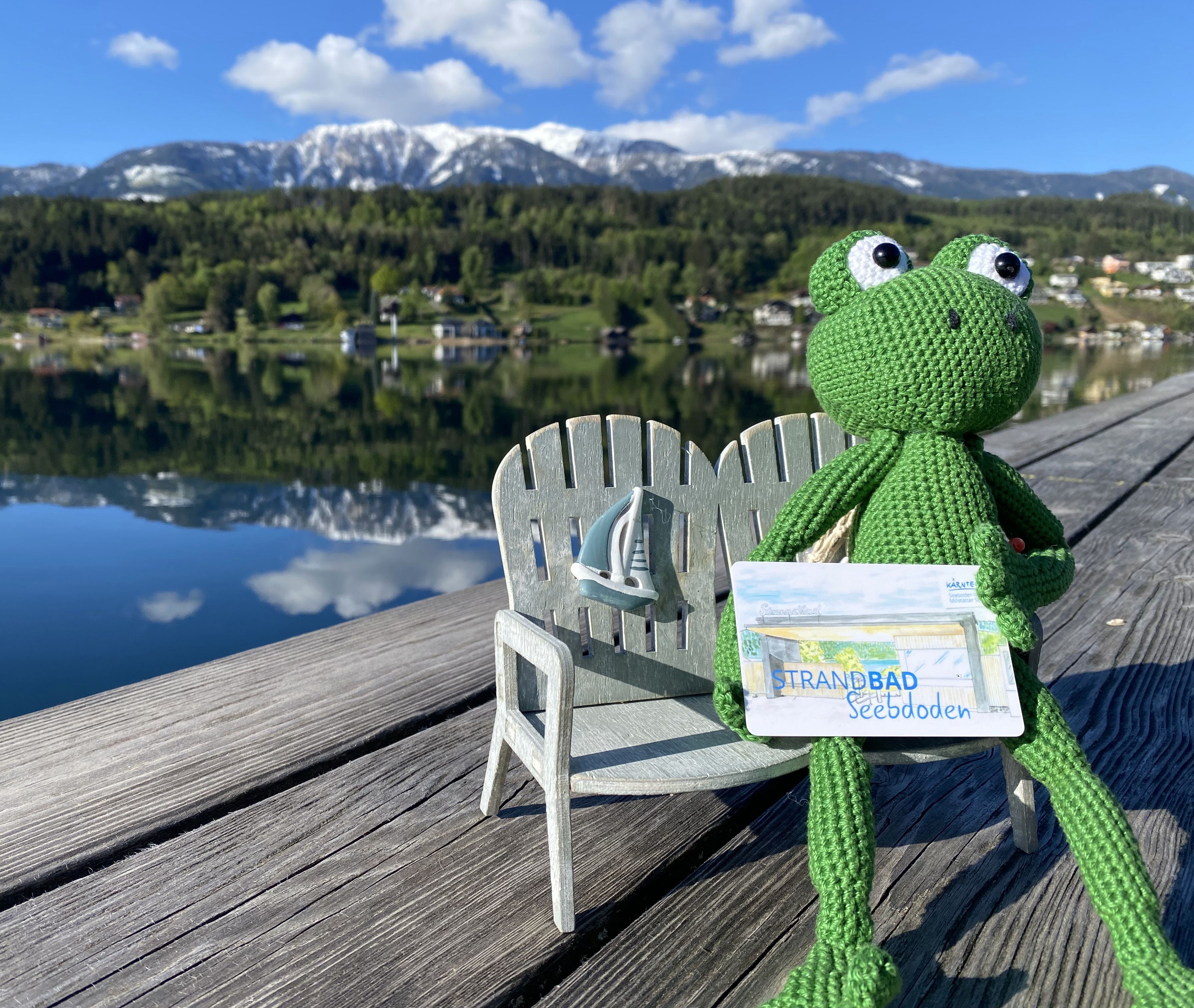 Das Maskottchen vom Strandbad Seeboden - ein kleiner gehäckelter grüner Frosch -  sitzt auf einen Miniaturbank am Steg des Strandbads. im Hintergrund sieht man den See und die Berge. 