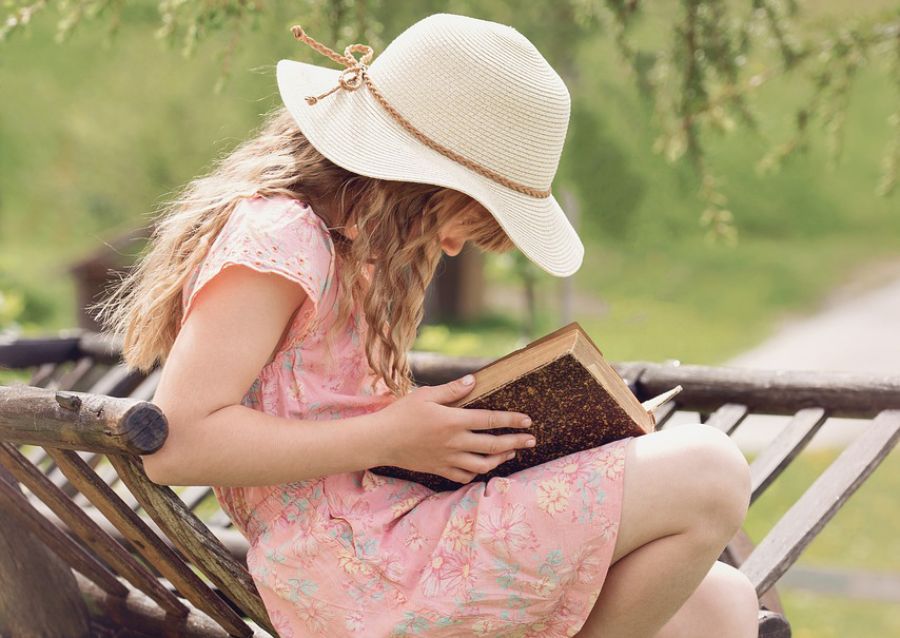 Ein Mädchen mit Blonden Haaren sitzt im freien und löießt ein Buch. Sie trägt einen Hut und hat blonde Haare. 