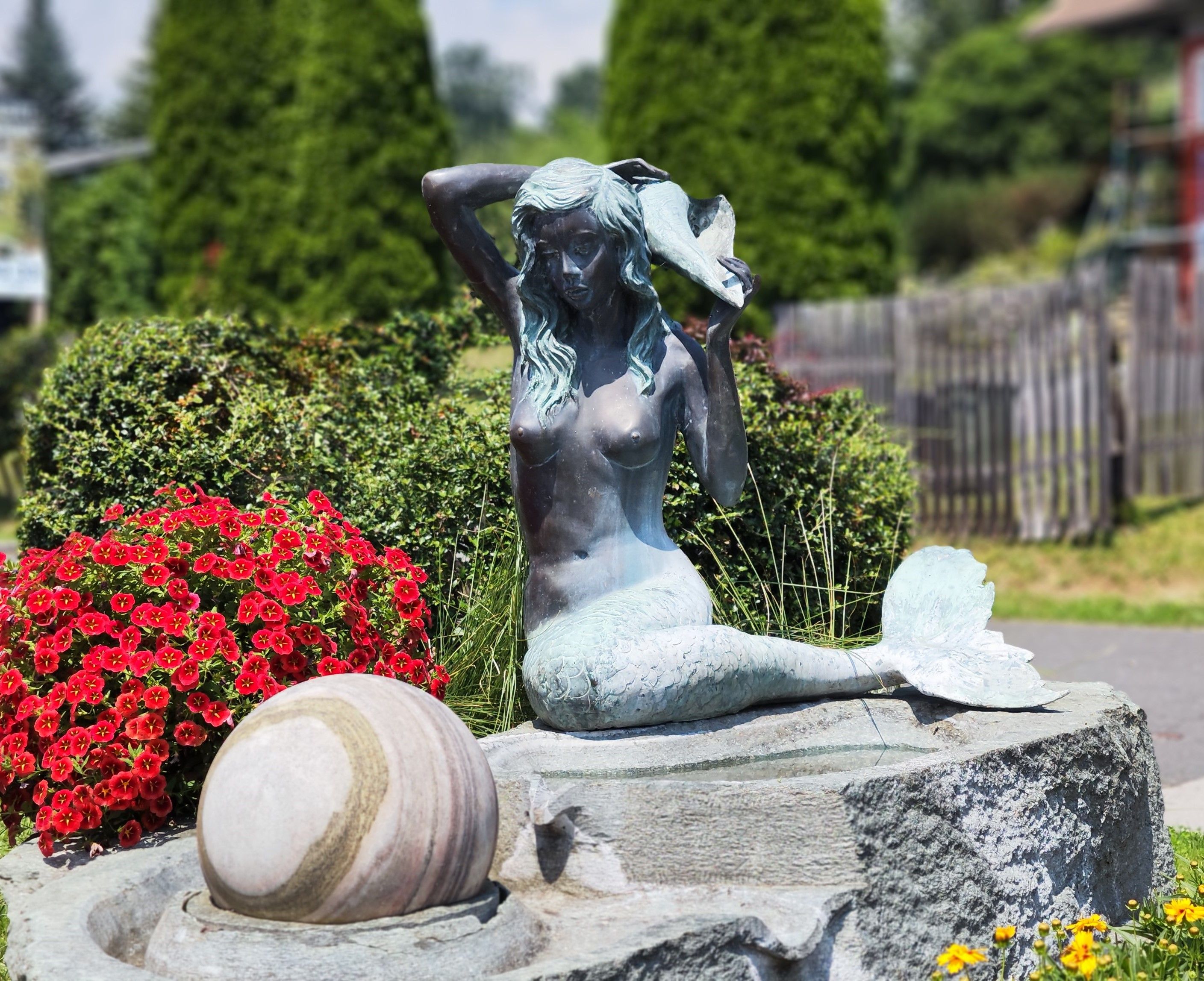 Auf dem Bild sehen sie die Seebodennixe als Statue auf einem Stein, nebenan stehen rote und gelbe Blumen.