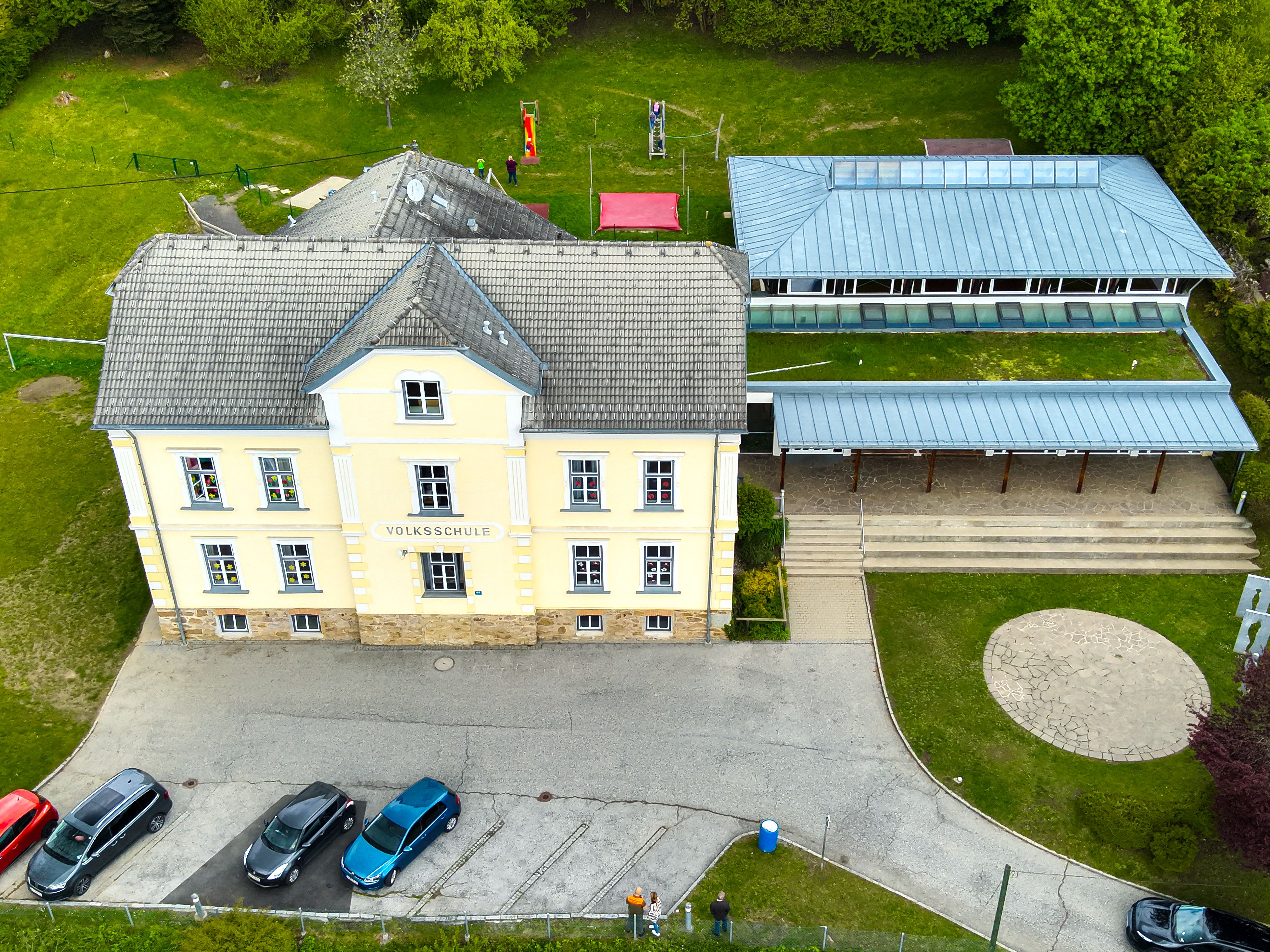 Die Volksschule Lieserhofen ist ein älteres Gebäude. Die Wände sind gelb mit der Aufschrift Volksschule. im hineren Bereich steht ein Spielplatz zur verfügung. 