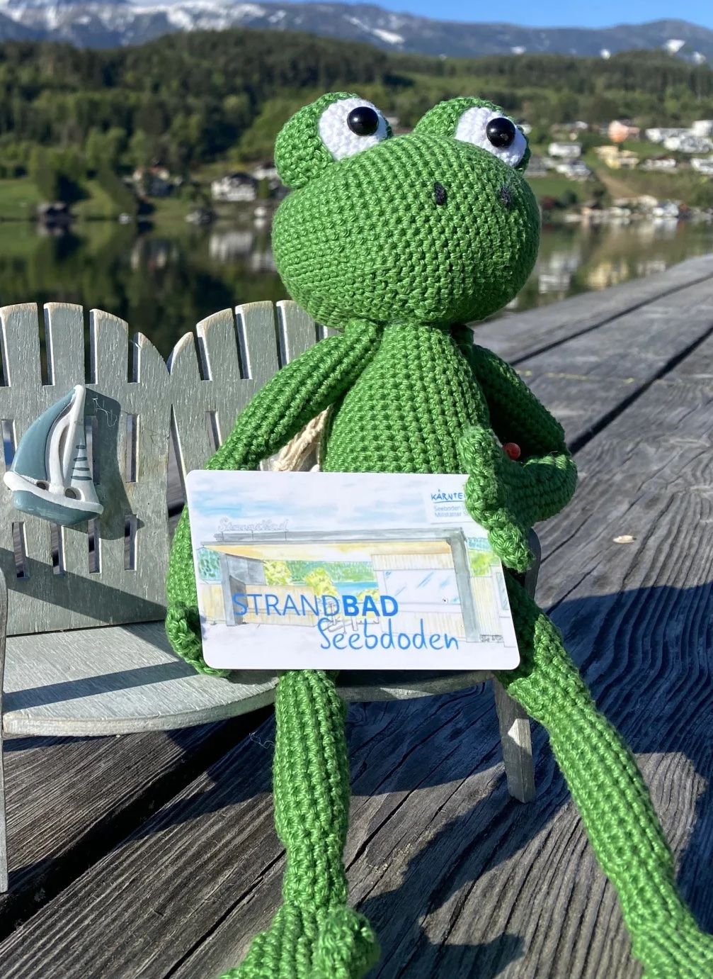 Das Maskottchen vom Strandbad Seeboden - ein kleiner gehäckelter grüner Frosch -  sitzt auf einen Miniaturbank am Steg des Strandbads. im Hintergrund sieht man den See und die Berge. 