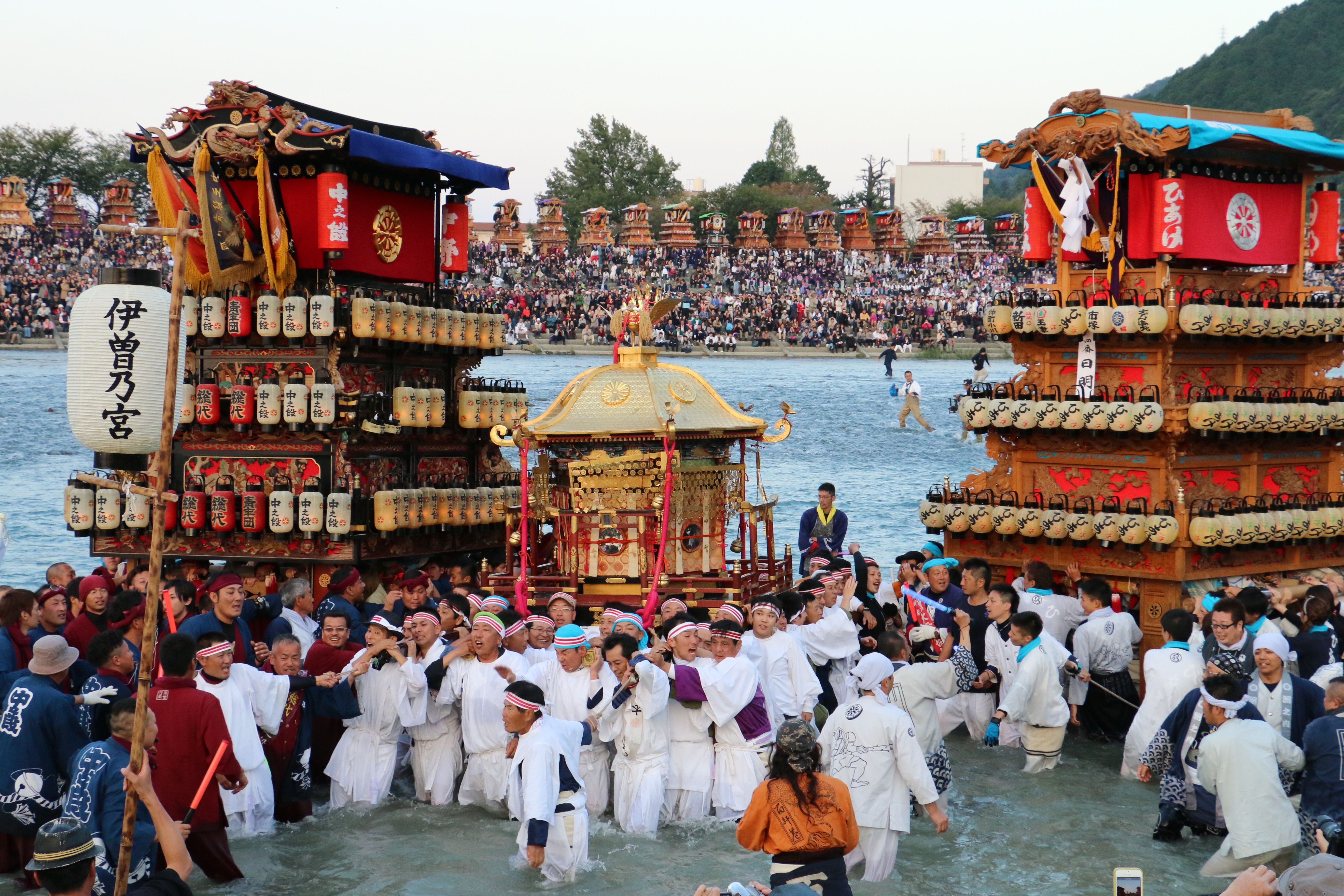 Saijō – die japanische Partnerstadt von Seeboden – Sie sehen ein Bild von einem japanischen Festival mit vielen Leuten die zwischen zwei Türmen am Wasser feiern.